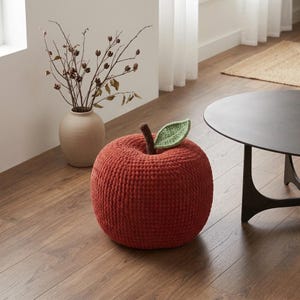 May include: A round, rust-colored, crocheted ottoman shaped like an apple. The top features a short, dark brown crocheted stem and a single, textured light green crocheted leaf attached to the center of the rounded, soft-textured surface.