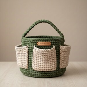 May include: A handmade, olive green crochet basket with a handle and three beige pockets. The basket has a round shape and a small, rectangular tag that reads "CrochetNestl". The crochet work is visible and detailed.