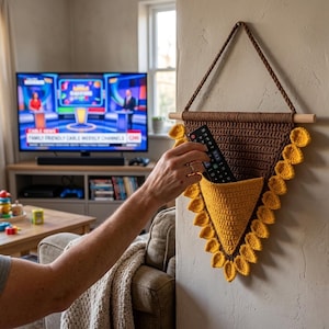 May include: A triangular wall-hanging organizer made of brown and yellow crochet yarn. The piece features a wooden dowel at the top with a braided cord for hanging and a yellow pocket containing a black remote control, accented by yellow petals.