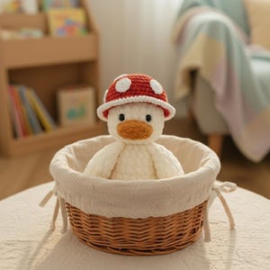 May include: A handmade white plush duck wearing a red mushroom hat with white spots, sitting in a woven basket with a cream-colored liner. The duck has an orange beak and black eyes. The basket is on a light-colored surface.