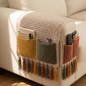 K&ouml;nnte beinhalten: Ein beigefarbener H&auml;kel-Sofa-Armlehnen-Organizer mit drei Taschen in Senfgelb, Olivgr&uuml;n und Rost. Die Taschen enthalten B&uuml;cher, Stifte, Brillen und Fernbedienungen. Der untere Rand hat Quasten in passenden Farben.