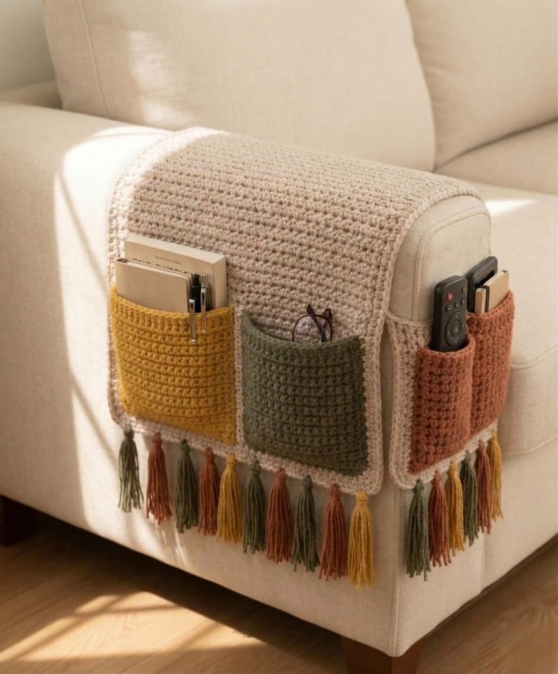 K&ouml;nnte beinhalten: Ein beigefarbener H&auml;kel-Sofa-Armlehnen-Organizer mit drei Taschen in Senfgelb, Olivgr&uuml;n und Rost. Die Taschen enthalten B&uuml;cher, Stifte, Brillen und Fernbedienungen. Der untere Rand hat Quasten in passenden Farben.