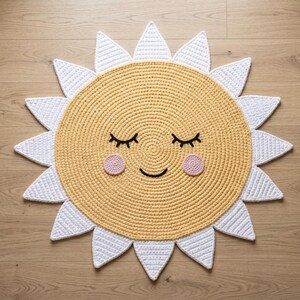 Puede incluir: Una alfombra con forma de sol, tejida a ganchillo en amarillo y blanco, con una cara sonriente. La alfombra tiene un centro amarillo con rayos blancos, ojos cerrados y mejillas rosas. Ideal para una habitaci&oacute;n infantil, esta alfombra a&ntilde;ade un toque alegre.