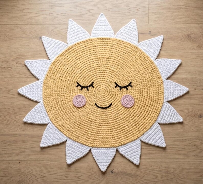 Puede incluir: Una alfombra con forma de sol, tejida a ganchillo en amarillo y blanco, con una cara sonriente. La alfombra tiene un centro amarillo con rayos blancos, ojos cerrados y mejillas rosas. Ideal para una habitaci&oacute;n infantil, esta alfombra a&ntilde;ade un toque alegre.