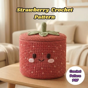 May include: A red crocheted container shaped like a strawberry, with a green stem and leaves on the lid. The container has white speckles and a smiling face. The image includes the text "Strawberry Crochet Pattern" and "Crochet Pattern PDF".