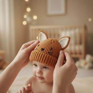 Pode incluir: Um chap&eacute;u de beb&ecirc; de malha em uma cor marrom quente, com orelhas de raposa com detalhes internos brancos e tra&ccedil;os faciais bordados. O chap&eacute;u est&aacute; sendo gentilmente colocado na cabe&ccedil;a de um beb&ecirc;. O fundo &eacute; suavemente iluminado com tons quentes.