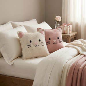 Peut inclure: Deux coussins carr&eacute;s en forme de chat sur un lit. L'un est cr&egrave;me avec des accents roses, l'autre est rose. Les deux ont des moustaches, des yeux et un nez noirs brod&eacute;s. Les coussins sont sur un lit avec des draps blancs et une couverture rose.
