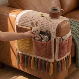 K&ouml;nnte beinhalten: Ein geh&auml;kelter Sofalehnen-Organizer in Beige, Gelb und Rostt&ouml;nen. Er hat mehrere Taschen zur Aufbewahrung, eine Quastenfranse und einen kleinen Stoffhasen. Eine Tasse und ein L&ouml;ffel liegen obenauf.
