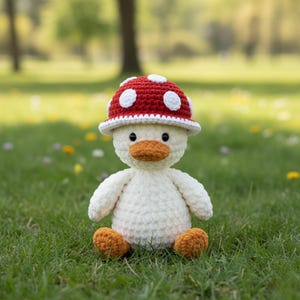 May include: A handmade, crocheted duck plushie with a red mushroom cap featuring white polka dots. The duck has a white body, orange feet and beak, and black eyes. It is sitting on green grass.