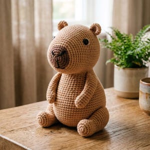 Pu&ograve; includere: Un giocattolo di peluche di capibara all'uncinetto fatto a mano, nei toni del beige e del marrone. L'animale di peluche ha un corpo arrotondato, piccole orecchie e un muso marrone scuro. &Egrave; seduto su una superficie di legno.