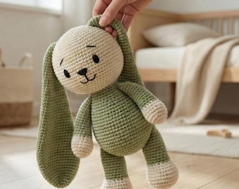 May include: A handmade crochet bunny plush toy. The bunny has long, floppy ears, a cream-colored head, and a green body with cream-colored accents on the feet and hands. The bunny has black eyes and a stitched nose and mouth.