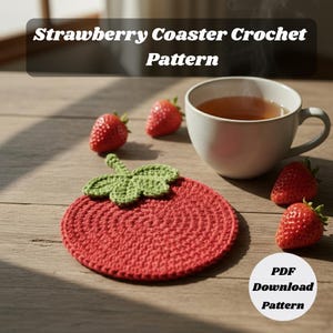 Puede incluir: Un posavasos de fresa rojo tejido a ganchillo con hojas verdes, junto a fresas frescas y una taza de té. El texto "Strawberry Coaster Crochet Pattern" está en la parte superior. Hay un patrón de descarga en PDF disponible.