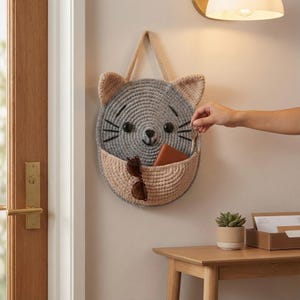 K&ouml;nnte beinhalten: Ein geh&auml;kelter Wandorganizer in Katzenform in Grau und Beige mit einer Tasche zur Aufbewahrung. Die Katze hat schwarze Schnurrhaare und Augen sowie beige Ohren. Eine Person legt Schl&uuml;ssel in die Tasche. Der Organizer h&auml;ngt an einer Wand.