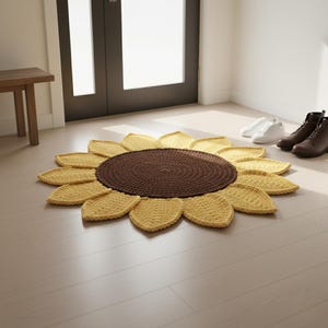 May include: A handmade, crochet sunflower rug with a brown center and yellow petals. The rug is placed on a light-colored wooden floor near a door and a small wooden bench. The rug's diameter is approximately 36 inches.