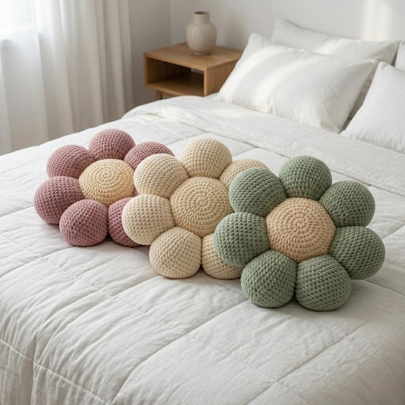 May include: Three handmade crochet flower pillows in shades of pink, cream, and green rest on a white bedspread. The pillows are arranged in a flower shape, with each petal made of multiple crocheted spheres. The bed is in a well-lit room.