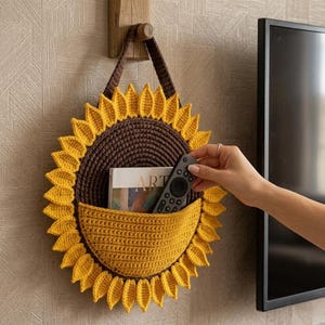 May include: A round, crocheted sunflower wall organizer featuring a dark brown center and bright yellow petals. It includes a yellow pocket containing a book titled ART and a black remote control, suspended by a brown crocheted strap.