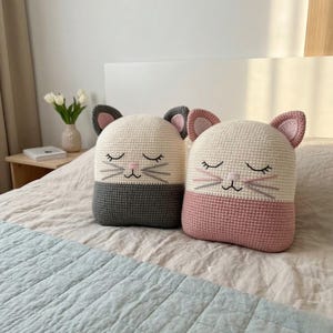May include: Two handmade cat-shaped pillows. The pillows are crocheted with a white top and a gray or pink bottom. Each pillow has a cat face with closed eyes, whiskers, and a pink nose. The pillows are on a bed.