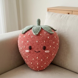 Könnte beinhalten: Ein handgefertigtes, gehäkeltes Erdbeer-Kissen mit einem lächelnden Gesicht. Das Kissen ist hauptsächlich rot-rosa mit weißen Samendetails und grünen Blättern. Das Kissen hat einen Durchmesser von etwa 30 cm.