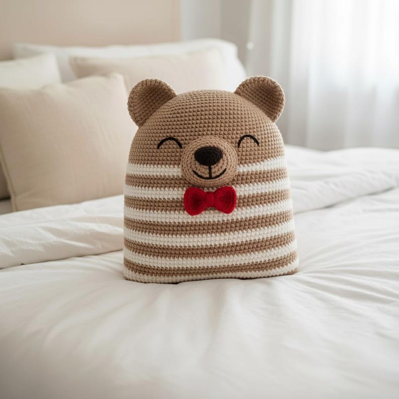 May include: A handmade crochet bear-shaped pillow with a smiling face and a red bow tie. The pillow features brown and white horizontal stripes and is placed on a white bed. The bear has small, rounded ears.