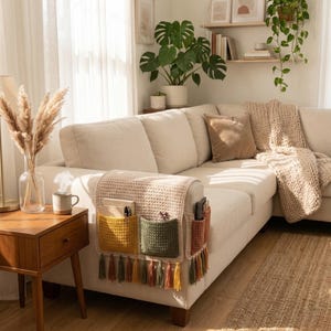 K&ouml;nnte beinhalten: Beiges Ecksofa mit einem geh&auml;kelten Armlehnen-Organizer. Der Organizer hat drei Taschen in Gelb, Gr&uuml;n und Rost, jede mit Quasten. Eine beige Decke und ein brauner Teppich sind ebenfalls zu sehen.