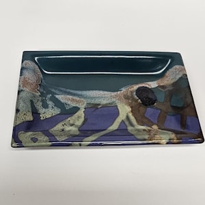 May include: Rectangular ceramic tray with a dark teal interior and a decorative exterior. The exterior features a blend of purple, brown, and cream-colored abstract designs. The tray is suitable for serving or display.