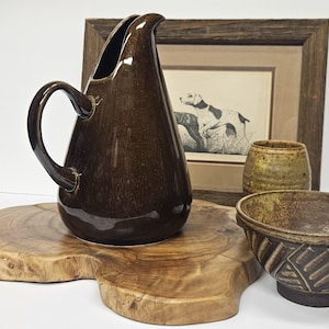May include: A brown ceramic pitcher with a curved handle sits on a wooden slab. A framed print of a dog and two ceramic bowls are also visible. The pitcher has a glossy finish.