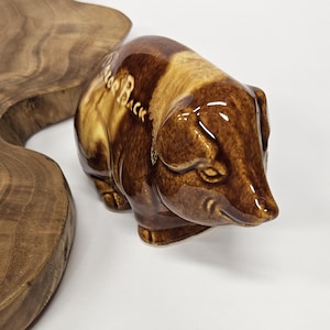 May include: A ceramic pig figurine in shades of brown and tan, with the words "For Back" on its side. The pig is glossy and has a detailed snout and ears. The figurine is on a white surface.