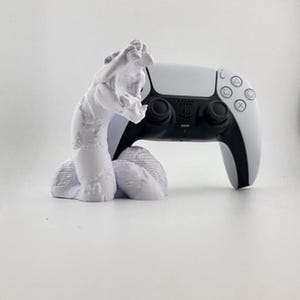 Puede incluir: Un mando blanco de PlayStation 5 descansa en un soporte blanco con forma de serpiente. El mando es blanco con detalles y botones negros. El soporte de la serpiente es una escultura detallada.