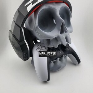 以下が含まれることがあります： グレーの頭蓋骨型ヘッドホンスタンド。黒と白のヘッドセットが上に置かれています。白と黒のゲームコントローラーが頭蓋骨の口に保持されています。「MAD_POWER」の文字がコントローラーに表示されています。