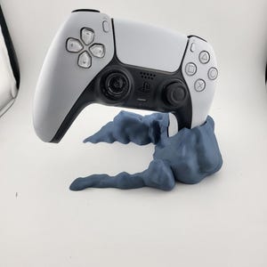 May include: A white and black PlayStation 5 controller resting on a dark blue cloud-shaped stand. The controller has a symmetrical design with various button shapes and a central touchpad. The stand provides support for the controller.