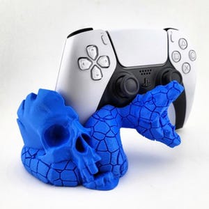 May include: A white and black PlayStation 5 controller rests against a blue skull-shaped holder with a snake design. The controller has a symmetrical design with buttons and a touchpad. The holder is a vibrant blue color.