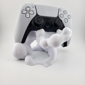 Puede incluir: Un mando blanco y negro de PlayStation 5 apoyado en un soporte blanco de forma abstracta. El mando presenta un diseño simétrico con varias formas de botones y un panel táctil central. El soporte tiene una forma única y fluida.