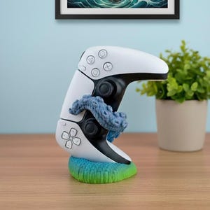 以下が含まれることがあります： 青い編み物のタコが巻き付いた白と黒のゲームコントローラー。 コントローラーは緑と青の編み物ベースの上にあります。 額入りの写真と鉢植えの植物が背景にあります。