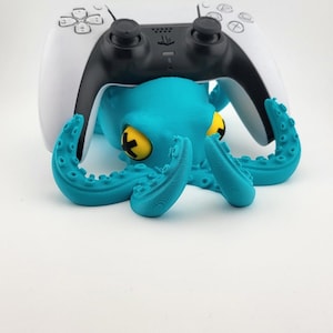 Op de afbeelding: Een turquoise octopusvormige controllerhouder met gele ogen en een zwart-witte gamecontroller die erop rust. De tentakels van de octopus zijn omhoog gebogen en ondersteunen de controller.