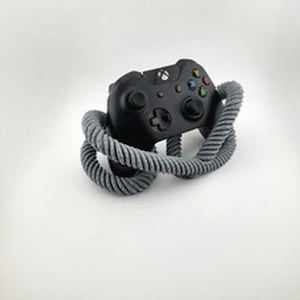 Op de afbeelding: Een zwarte Xbox-controller met gekleurde knoppen en een directioneel pad is omcirkeld door een grijs, gedraaid touw. De controller heeft twee joysticks en een centrale Xbox-knop. De achtergrond is effen wit.