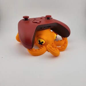 Puede incluir: Un mando de videojuegos rojo descansa sobre un soporte con forma de pulpo naranja. El pulpo tiene ojos amarillos y un diseño detallado de tentáculos. El mando tiene dos joysticks y varios botones.