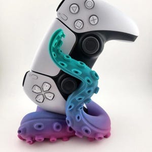 May include: A white and black game controller is held by a colorful octopus-shaped stand. The stand transitions from purple to blue to teal. The controller has buttons with symbols.