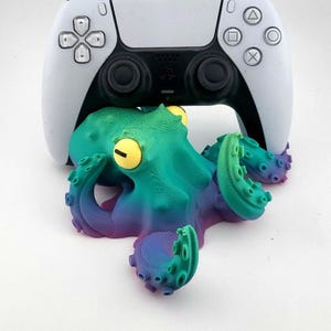 May include: A white PlayStation 5 controller rests atop a colorful, 3D-printed octopus figurine. The octopus features a gradient of green, blue, and purple hues, with yellow eyes. The controller's buttons and directional pad are visible.