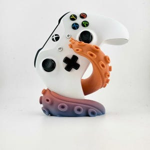 May include: A white Xbox controller is held by an orange, purple, and blue tentacle-shaped stand. The controller has black buttons and colored action buttons. The stand is designed to hold the controller upright.