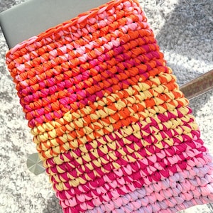 Könnte beinhalten: Eine farbenfrohe, handgehäkelte Laptoptasche. Die Tasche zeigt einen lebendigen Farbverlauf aus Orange, Pink und Gelb. Das strukturierte Design ist mit dickem Garn gefertigt und schafft ein einzigartiges und stilvolles Accessoire.