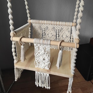 May include: A handmade macramé baby swing. The swing has a natural wood seat and a decorative macramé back and front. It is suspended by thick, twisted rope and includes wooden rings and tassels. The colour is off-white.