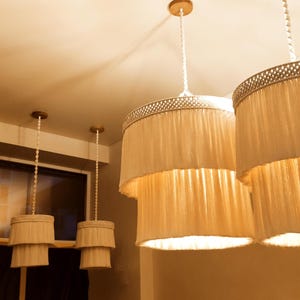 Puede incluir: Varias lámparas colgantes de color crema cuelgan del techo. Las pantallas tienen un diseño escalonado con una tela plisada y texturizada. Las luces están suspendidas por un cable blanco y tienen un adorno decorativo. Tema: decoración del hogar.