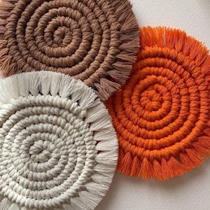 May include: Close-up of three handmade macrame coasters in shades of brown, orange, and white. Each coaster features a spiral design with a fringed edge. The coasters are made of thick, textured cotton cord.