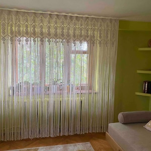 May include: Cream-colored macrame curtains hanging in front of a window. The curtains have a geometric pattern at the top and vertical strands that hang down. The window is framed by a wooden frame. The room has green walls and a couch.