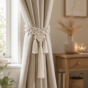 Könnte beinhalten: Beige Leinen-Vorhänge, gehalten von einem cremefarbenen Makramee-Vorhanghalter mit drei Quasten. Die Vorhänge hängen vor einem Fenster, mit einem hölzernen Beistelltisch und gerahmten Kunstwerken im Hintergrund.
