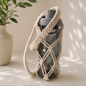May include: A dark gray plastic water bottle encased in a beige macrame holder with a braided shoulder strap. The bottle features a screw-top lid and a textured grip area. The holder is crafted from thick cord in a diamond-shaped net pattern.