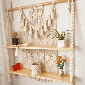 May include: A two-tiered macrame hanging shelf. The wooden shelves are suspended by woven rope and adorned with a macrame panel and tassels. The shelves display potted plants, candles, and decorative vases.