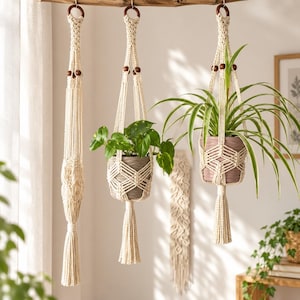 Può includere: Tre portapiante in macramè color bianco sporco appesi a un bastone di legno. Due portapiante contengono piante verdi in vaso, mentre uno è vuoto. Ogni portapiante presenta complessi motivi a nodi, perline di legno scuro e una lunga nappa.