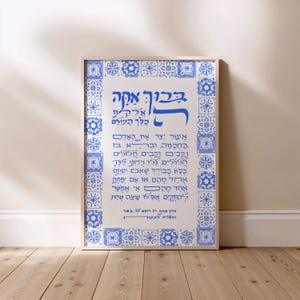 May include: Framed art print featuring Hebrew calligraphy in blue on a white background. The text is surrounded by a border of blue and white geometric tile patterns. The artwork is displayed on a wooden floor.