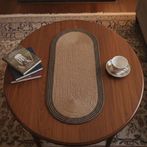 Peut inclure: Une table basse ronde en bois avec un tapis ovale tissé, des livres et une tasse à thé. Le tapis est beige avec une bordure marron foncé. Les livres sont empilés et la tasse à thé a un motif floral.